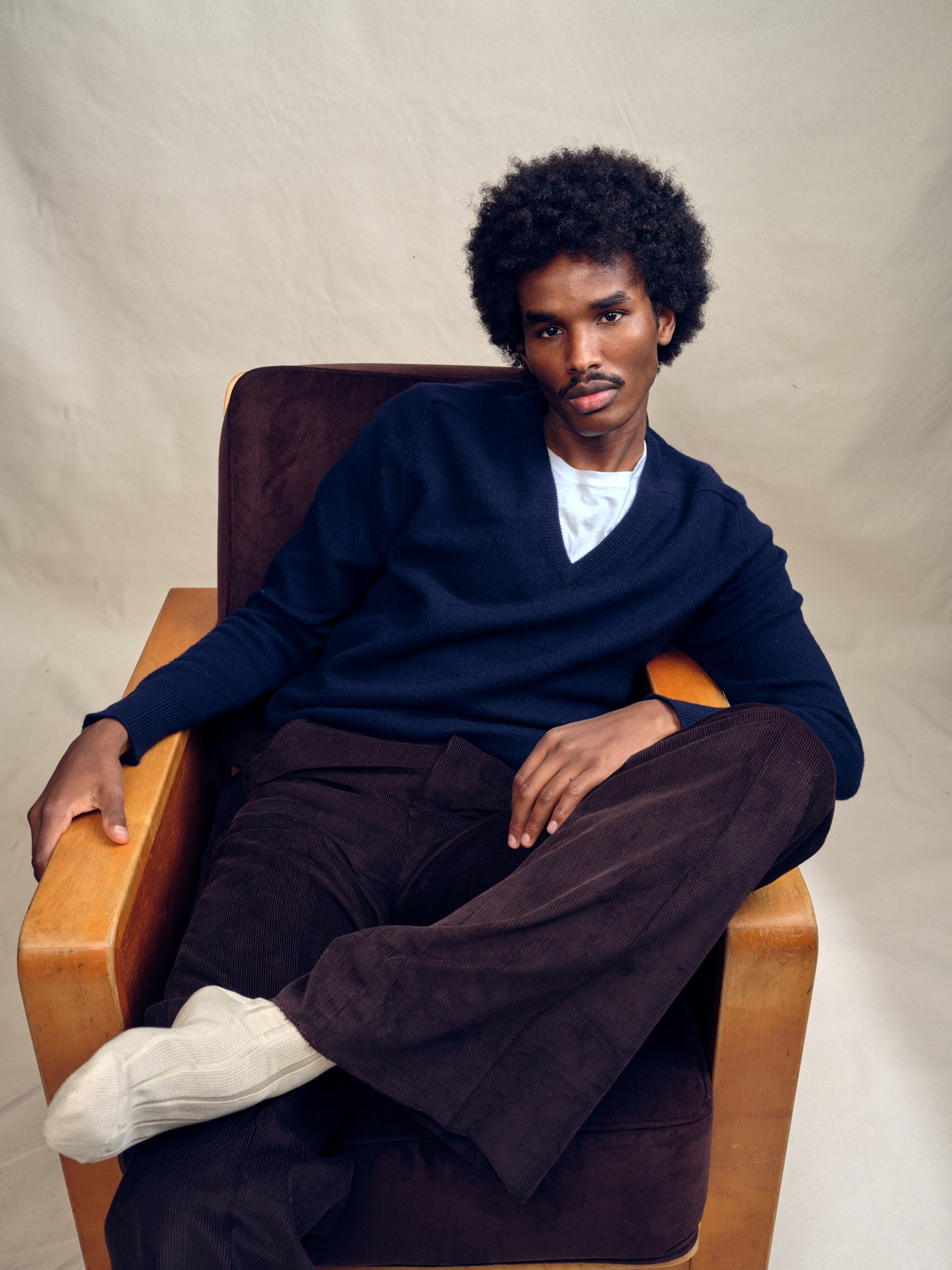 Man sitting in a chair wearing a navy Vintage Saks Fifth Avenue blue v-neck sweater, cashmere, made in Scotland and dark pants against a beige background
