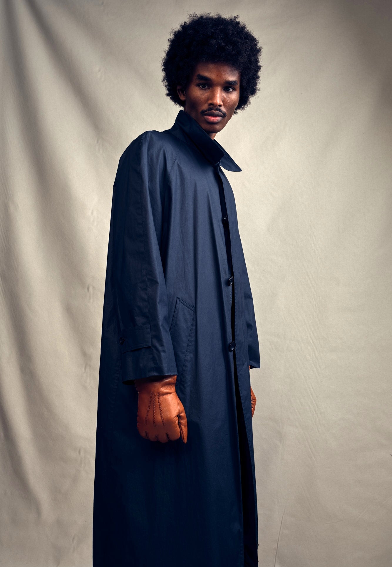 Man wearing a long navy Turnbull &Asser car coat in cotton and orange gloves against a plain background